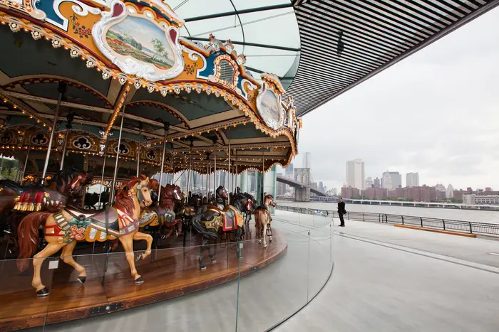 A photograph of a carousel with horses, with the East River seen on the right side